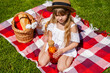 Cute little girl sitting on a blanket with lemonade in a glass bottle during a picnic in the park on the grass on a sunny summer day.