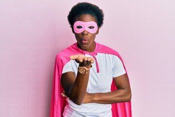 Wall Mural - Young african american girl wearing superhero mask and cape costume looking at the camera blowing a kiss with hand on air being lovely and sexy. love expression.