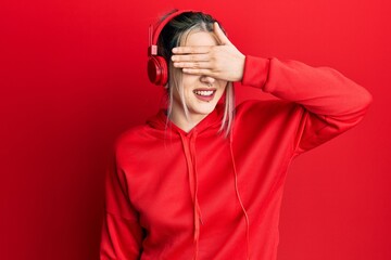 Wall Mural - Young modern girl wearing gym clothes and using headphones smiling and laughing with hand on face covering eyes for surprise. blind concept.