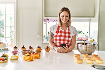 Poster - Young beautiful blonde woman wearing apron cooking pastries looking for recipe on smartphone looking positive and happy standing and smiling with a confident smile showing teeth