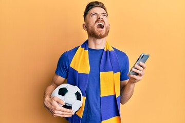 Wall Mural - Young redhead man wearing hooligan scarf and using smartphone angry and mad screaming frustrated and furious, shouting with anger looking up.