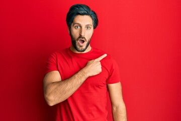 Young hispanic man wearing casual red t shirt surprised pointing with finger to the side, open mouth amazed expression.