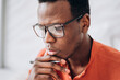 Thoughtful young African-American man with elegant glasses and pen wearing bright orange clothes in light room close view