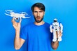 Young hispanic man holding drone and robot toy in shock face, looking skeptical and sarcastic, surprised with open mouth