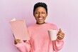 Young african american girl reading a book and drinking a cup of coffee smiling and laughing hard out loud because funny crazy joke.