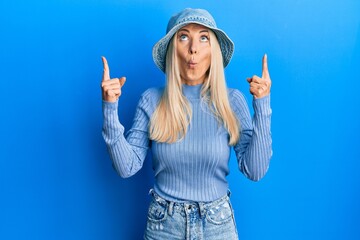 Poster - Young blonde woman wearing casual denim hat amazed and surprised looking up and pointing with fingers and raised arms.