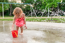 Little Child Playing With Water And Toys At Splash Pad In The Local Public Park During Hot Summer Day. Small Beautiful Girl In Pink Dress Having Fun At Fountain Playground.