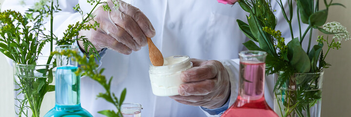 Concept of herbal natural organic ingredients in cosmetic. Young man scientist in white medical gown doing laboratory research and tests with plant and flower extracts creating face cream. Banner