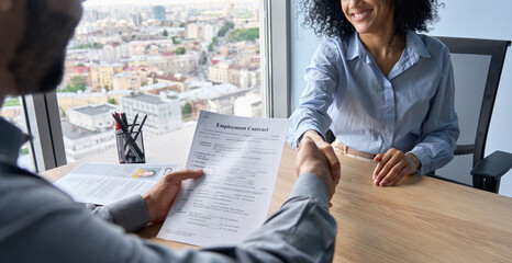 Wall Mural - Closeup of Indian latin ceo holding employment contract job offer hiring welcoming female African American newcomer worker manager shaking hands in contemporary office. Human resources concept.