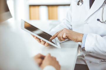 Unknown male doctor and patient woman discussing current health examination while sitting in clinic and using tablet computer. Perfect medical service in hospital. Medicine and healthcare concept