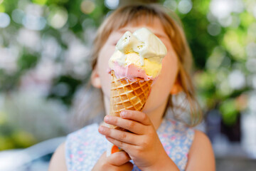 Little preschool girl eating ice cream in waffle cone on sunny summer day. Happy toddler child eat icecream dessert. Sweet food on hot warm summertime days. Bright light, colorful ice-cream