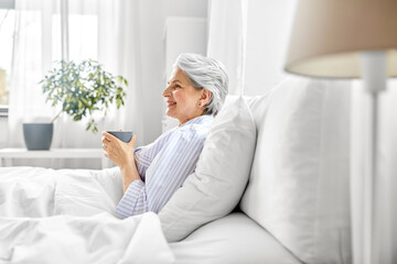 Poster - morning, old age and people concept - happy smiling senior woman in pajamas with cup of coffee sitting on bed at home bedroom