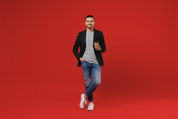 Wall Mural - Full length young smiling businessman latin man 20s with long hair ponytail wear black striped jacket grey shirt hold takeaway delivery craft paper brown cup coffee to go isolated on red background