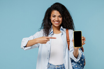 Young african american girl teen student in denim clothes backpack point finger on mobile phone blank screen workspace isolated on blue background Education in high school university college concept.