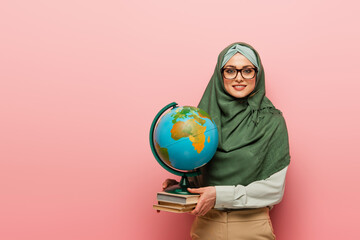 smiling muslim teacher with textbooks and globe looking at camera on pink background