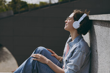 Wall Mural - Young side view smiling caucasian woman in jeans clothes headphones eyeglasses listen to music leaning on building wall sit on concrete steps outdoors walk look aside People urban lifestyle concept.