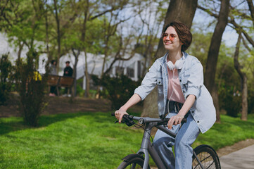 Side view young satisfied happy sporty woman in jeans clothes headphones riding bicycle bike on sidewalk in city spring park outdoors, look aside People active urban healthy lifestyle cycling concept