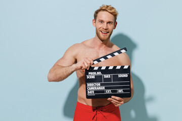 Handsome young fun sexy man in red shorts swimsuit relax near hotel pool holding classic black film making clapperboard isolated on pastel blue background. Summer vacation sea rest sun tan concept