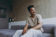 © ViDi Studio - Happy cheerful fun smiling friendly young african american man 20s wearing beige t-shirt sweatpants sitting on grey sofa indoors apartment procrastinate looking aside, rest on weekends stay at home