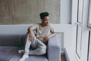 Full length pensive young african american man 20s with curly hair in beige t-shirt sweatpants sitting on comfortbale grey sofa indoors apartment looking at window resting on weekends staying at home