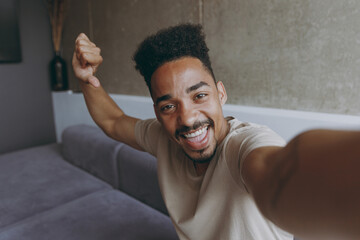 Close up confident young african american man in beige casual t-shirt sit on grey sofa indoors apartment doing selfie shot on mobile phone point thumb finger on himself rest on weekends stay at home