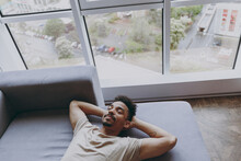 Top View From Above Relaxed Young African American Man 20s Wear Casual Beige T-shirt Sweatpants Lay Down On Grey Sofa Sleeping Hold Hand Behind Neck Indoors Apartment Rest On Weekends Staying At Home.