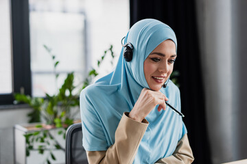 Wall Mural - positive arabian woman in hijab and headset holding pen while working in call center