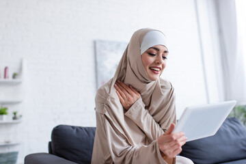 Wall Mural - cheerful arabian woman in hijab using digital tablet at home