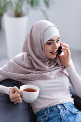 Wall Mural - young arabian woman in hijab talking on smartphone while holding cup of tea