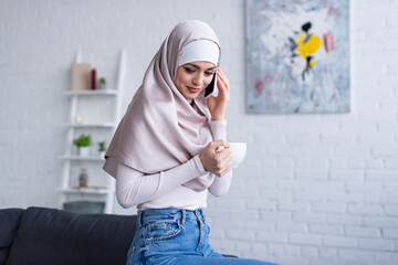 Wall Mural - smiling arabian woman holding cup of tea while talking on mobile phone