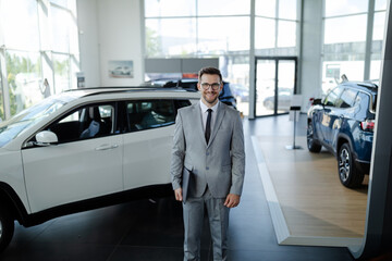 Wall Mural - Confident young car dealer standing in showroom and holding laptop computer while posing.