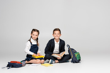 Wall Mural - happy schoolkids in prestigious uniform sitting with backpacks and books on grey.