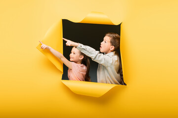 Wall Mural - kids pointing away with fingers through hole on yellow background.