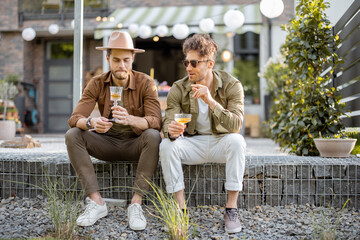 Wall Mural - Two male friends having a close conversation, sitting with drinks on a porch of the country house outdoors. Fun summer time in a country house, male friendship