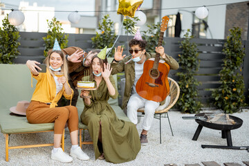 Wall Mural - Friends in face masks having a birthday party, making selfie with a cake or having video call on phone while sitting together at backyard outdoors