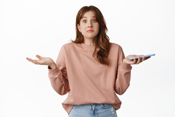 Wall Mural - Portrait of confused redhead woman shrugging shoulders, holding mobile phone and look clueless, cant understand, dont know nothing, standing over white background