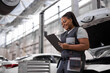 © Roman - black female Auto Mechanic Making Car Checkup, in Uniform Writing and holding Clipboard of Service Order Indoors. Repair service. Woman Car Repairing Concept