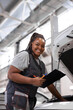 © Roman - Afro female Auto Mechanic Making Car Checkup, in Uniform Writing and holding Clipboard of Service Order Indoors. Repair service. Woman Car Repairing Concept. Black woman look at camera smiling