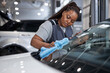 © Roman - Serious Female afro auto mechanic cleaner is cleaning car window glass with blue rag, side view on adorable black woman in car service, working alone, washing vehicle