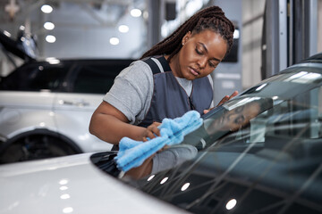 Wall Mural - Serious Female afro auto mechanic cleaner is cleaning car window glass with blue rag, side view on adorable black woman in car service, working alone, washing vehicle