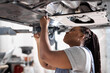 © Roman - Afro auto mechanic work in garage, car service technician woman in overalls check and repair customer car at automobile service center, inspecting car under body and suspension system