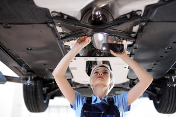 Wall Mural - The concept of feminism and women's equality. woman in overalls in auto service. short haired young caucasian woman shines a lantern at the bottom of the lifted car, concentrated and focused