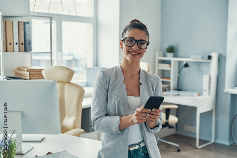 Beautiful young woman in smart casual using smart phone and smiling while standing in the office - obrazy, fototapety, plakaty 