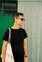 30 Year Old Man Standing Outdoors In Front Of A Green Door Holding A Linen Bag And Wearing Sunglasses