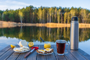 Wall Mural - Breakfast with hot tea, cheese sandwich and honey on wooden table in morning next to the lake. Nature and food concept