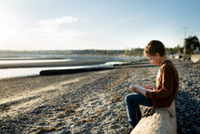 Boy Sits On Driftwood Log Reading Book At Beach