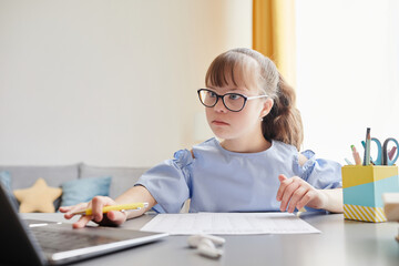 Wall Mural - Portrait of cute girl with down syndrome doing homework while sitting at desk in cozy interior and using laptop, copy space