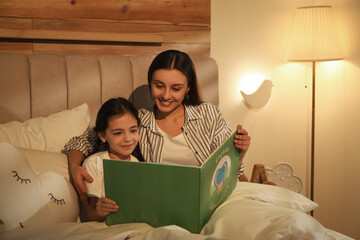 Poster - Little girl with mother reading book in bedroom lit by night lamp