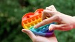 Colorful antistress sensory heart-shaped toy fidget push pop it on green nature background. Adult female hand playing rainbow toy pop it toy simple dimple. Anti Stress Sensory Toy. Trend 2021. 