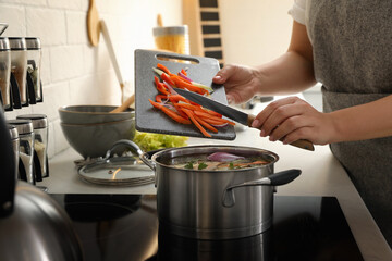 Wall Mural - Woman putting cut vegetables into pot with delicious bouillon on stove in kitchen, closeup